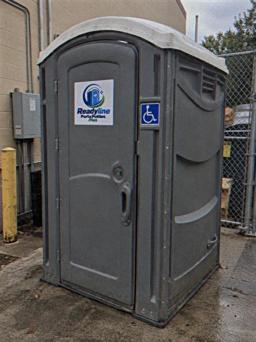 Grey ADA porta potty next to yellow bollards and a chain-link fence
