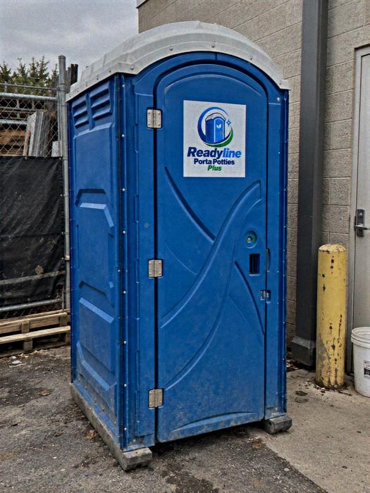 Blue porta potty against a building wall beside a yellow bollard