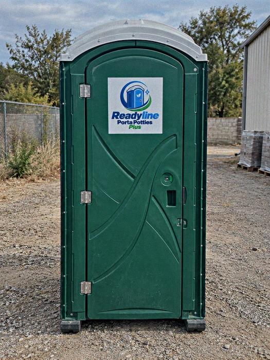 Green porta potty on a gravel lot with wooden pallets nearby