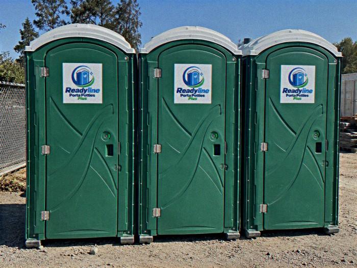 Three green porta potties lined up on a dirt and gravel job site