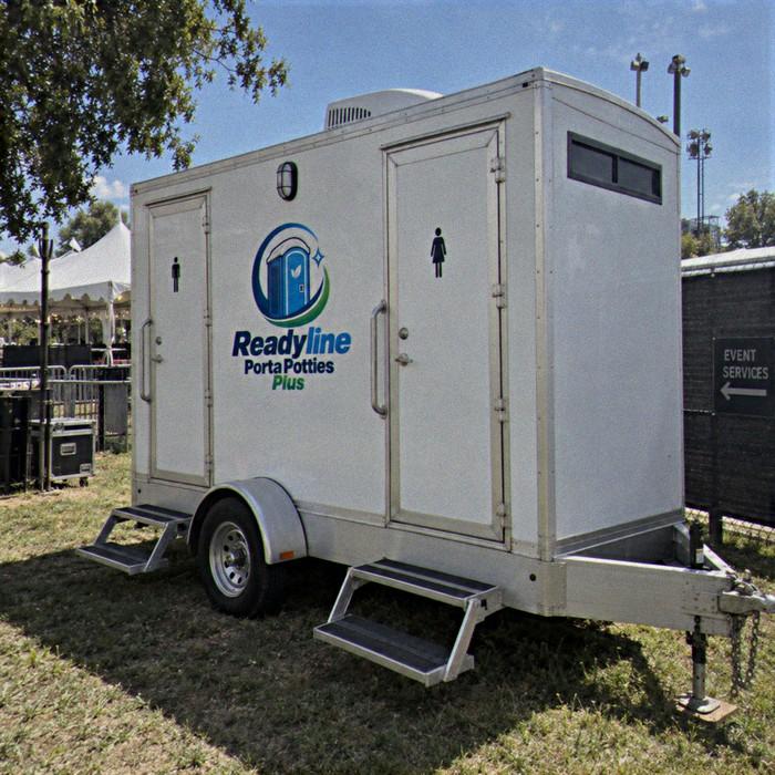 White restroom trailer on grass beside event tents and metal fencing under blue sky