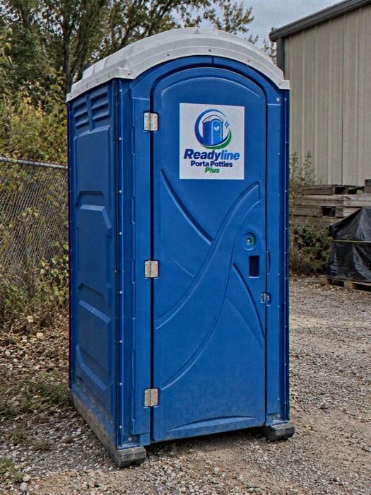 Blue portable toilet on gravel beside a metal shed and trailer