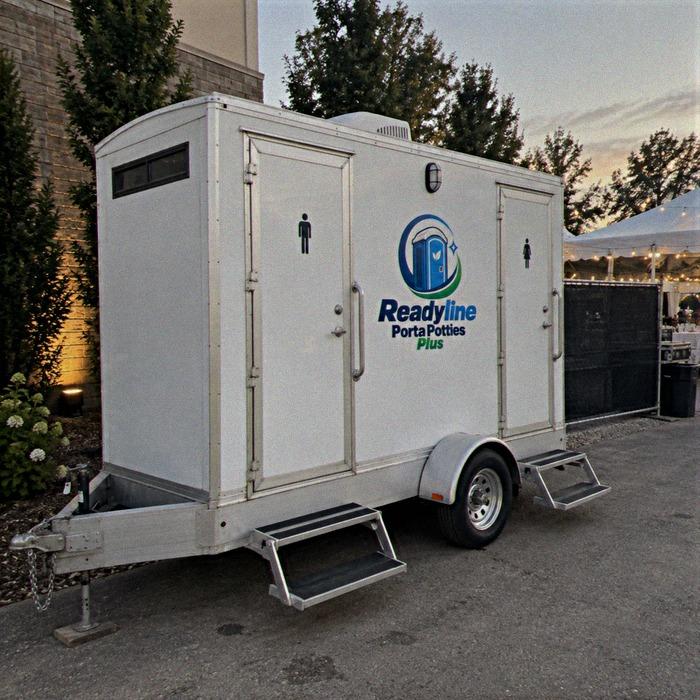 White restroom trailer at dusk with warm string lighting and trees around it