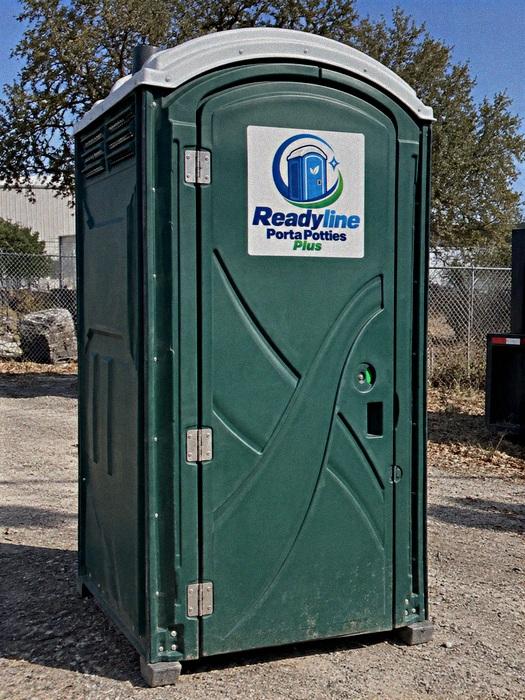 Green porta potty on gravel beside a chain-link fence and bushes