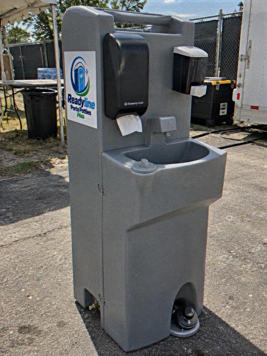 Grey hand-wash sink stand with foot pump visible, set on grass beside a fence