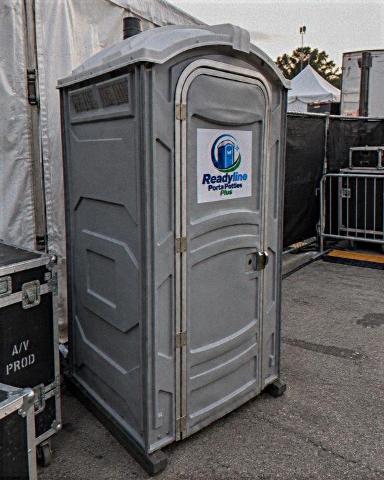 Grey single porta potty beside black road cases and metal barriers at an event