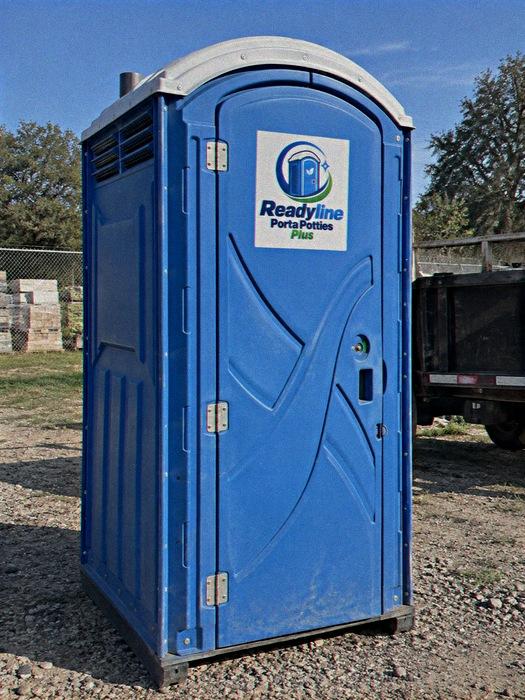 Blue portable toilet on gravel with a trailer and trees in the background