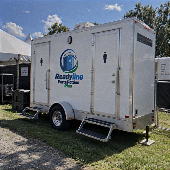 White restroom trailer on grass with white event tents behind and clear blue sky
