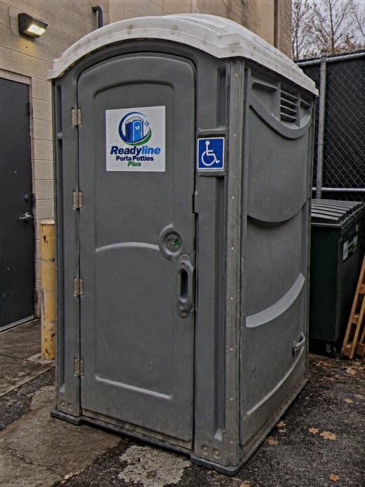 Grey wheelchair accessible porta potty beside a building and trash bins